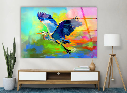 A vibrant, abstract background with bold strokes of blues, greens, yellows, and pinks complements the soaring heron in this glass printing.