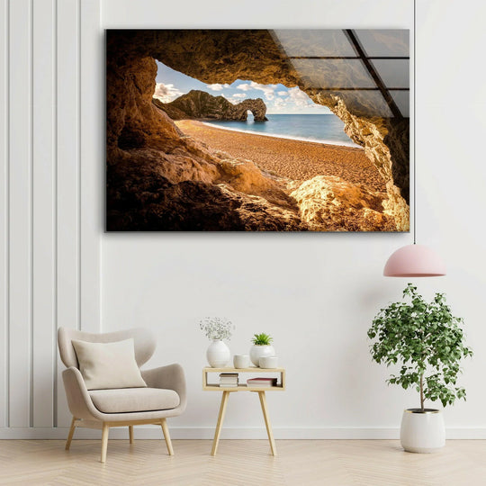 The stillness of the landscape captures a serene beach vista through a cave, showcasing a golden, coastal scene, amplified by its vibrant glass wall art decor.