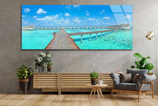 Tropical Maldives glass wall art showcases a long wooden pier and turquoise water and features a scenic landscape for modern wall decor above a sofa