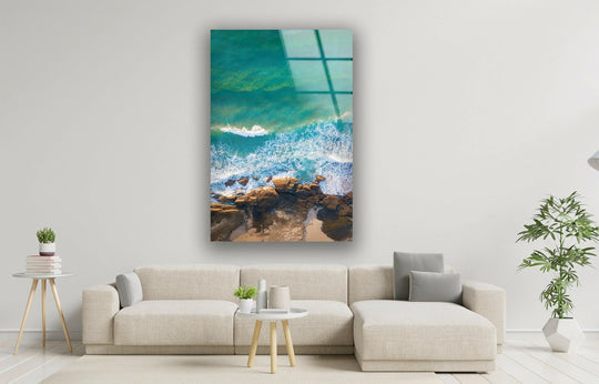 Glass wall art showcases the shimmering texture of aqua ocean currents and the rough texture of the rocky shoreline.