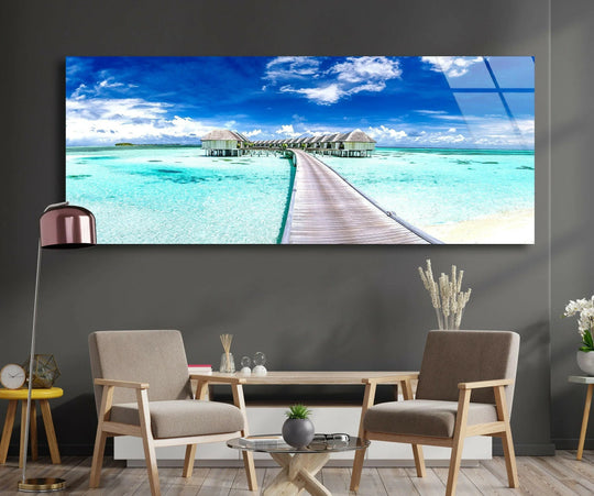 Maldives Beaches Landscape glass wall art showcases a scenic tropical view and a wooden walkway with turquoise water and a blue sky and white clouds above sofa
