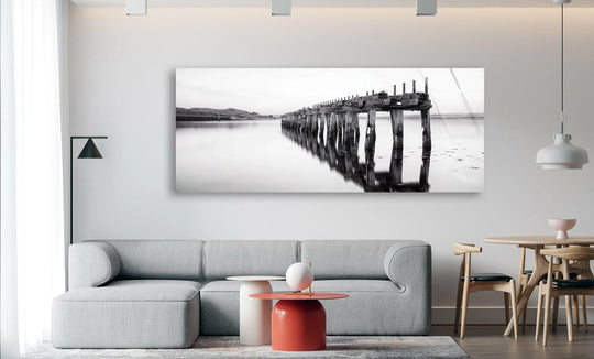 Old Wooden Railway Bridge glass wall art showcases a scenic landscape and panoramic view with a weathered wooden structure reflected in calm water above a sofa